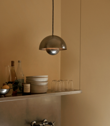 Lampa wisząca z polerowanego metalu nad półką z ceramicznymi misami i szklankami na tle piaskowej ściany.